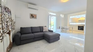 A modern living room with a grey sectional sofa, a white marble floor, and a kitchen in the background. - Loft 'Oltremare' Apartment with Private Terrace, Wi-Fi & Air Conditioning (Capoliveri)