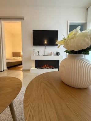 Ein modernes Wohnzimmer mit Kamin, einem an der Wand befestigten Flachbildfernseher und einer weißen Vase mit Blumen auf einem Holztisch.