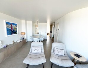 Ein modernes Hotelzimmer mit Essbereich, zwei Lounge-Sesseln mit Kissen, einem kleinen Tisch mit Fernbedienungen und einem Gemälde an der Wand.