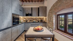 A modern kitchen with a stone wall, wooden ceiling, and a dining table with a fruit bowl. - Elegant Tuscan Home in Chianti Near Florence (Sant'Andrea in Percussina)