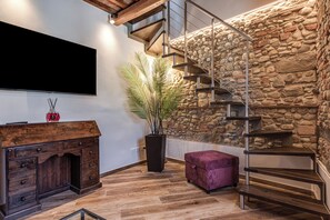 Ein modernes Interieur mit einem Holzkabinett, einer großen Kübelpflanze, einem violetten Hocker, einer Treppe an einer Steinwand und einem an der Wand montierten Flachbildfernseher.