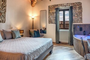 Un dormitorio con una cama grande, una mesita de noche, un espejo, una pared de piedra, una ventana y un baño con bañera.