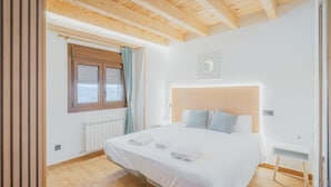 A bedroom with a wooden ceiling, a bed with white linens, a window with curtains, and a radiator.