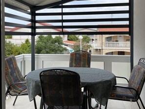 Ein überdachter Außenbereich mit Tisch und Stühlen, Blick auf Wohnhäuser und einem Balkongeländer.