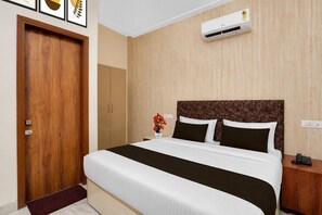 A hotel room with a bed, bedside table, nightstand, and an air conditioning unit.