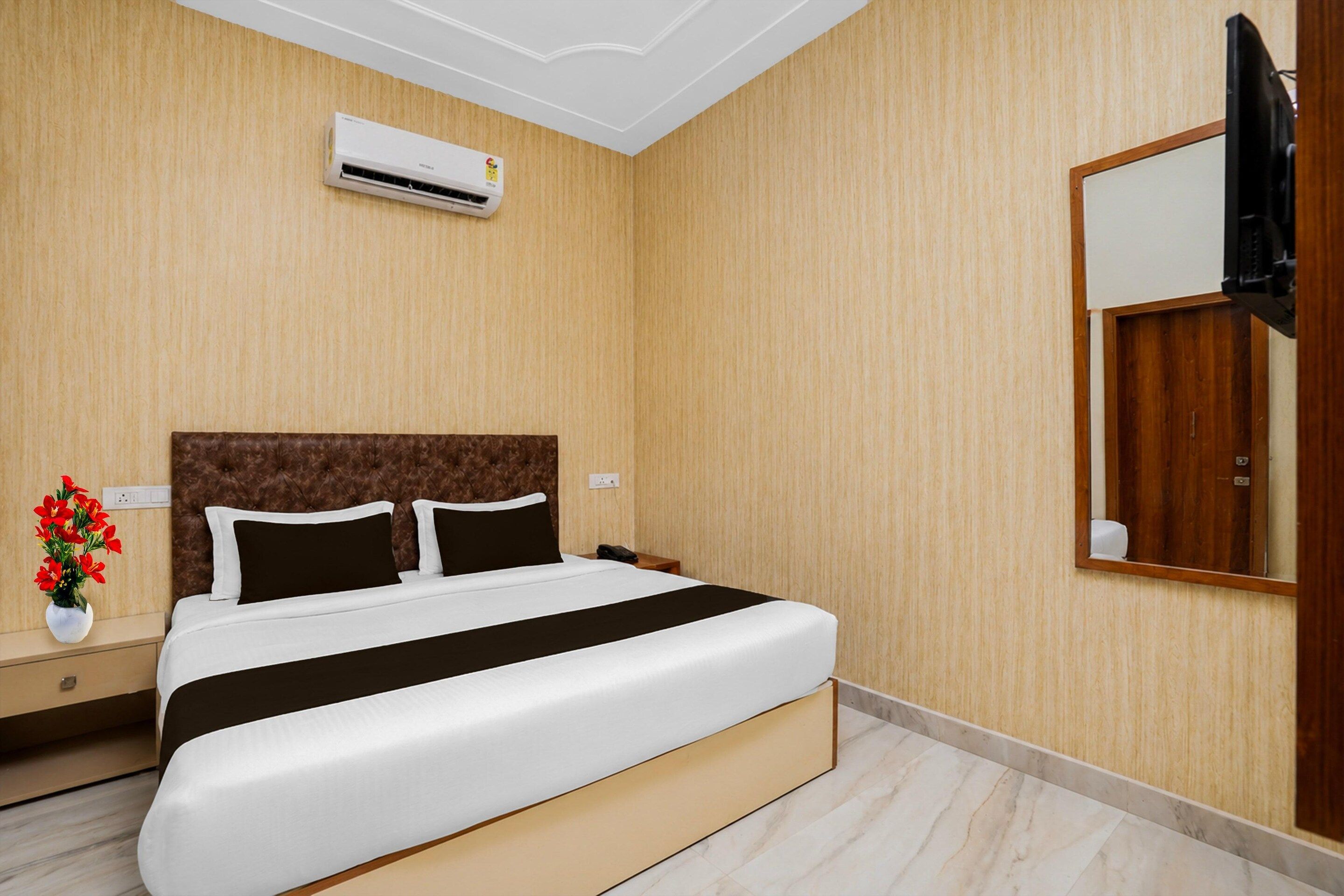 A hotel room with a bed, a nightstand, a vase with flowers, a wall-mounted TV, and an air conditioning unit.