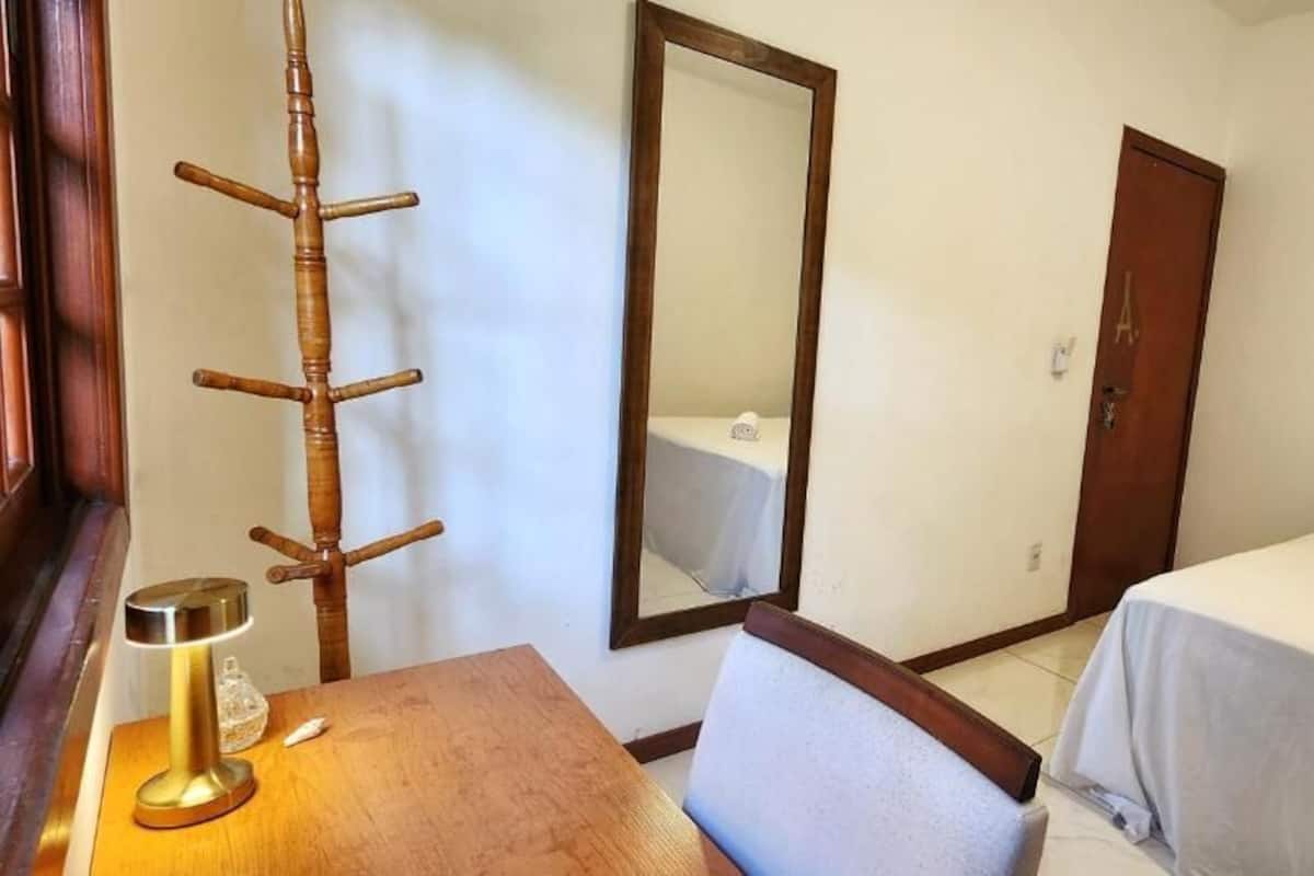 A hotel room with a wooden desk, a chair, a bed with a white bedsheet, a mirror, and a wooden door.