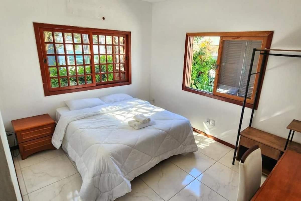 A bedroom with a bed, a wooden nightstand, a wooden desk, and a window with a view of greenery.