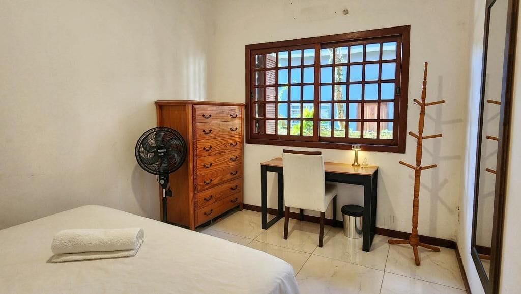 Un dormitorio con cama, cómoda de madera, escritorio con silla, ventilador de pie, ventana con vistas y una mesita de noche con lámpara.