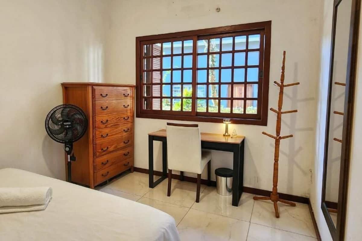 A bedroom with a bed, a wooden dresser, a desk with a chair, a standing fan, a window with a view, and a small table with a lamp.