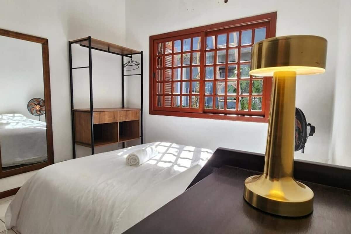 A room with a bed, a wooden shelf, a mirror, a window with a grid pattern, and a table lamp.
