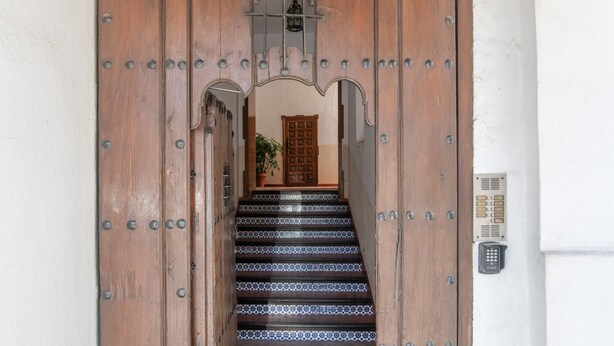 Una puerta de madera con refuerzos metálicos, una escalera de baldosas con diseño y un portero automático en la pared.