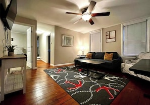 Un salon avec un ventilateur de plafond, un canapé, une table basse et un tapis à motifs.
