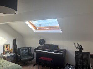 Een kamer met dakraam, een piano, een stoel, een klein tafeltje met een afbeelding en een speaker.