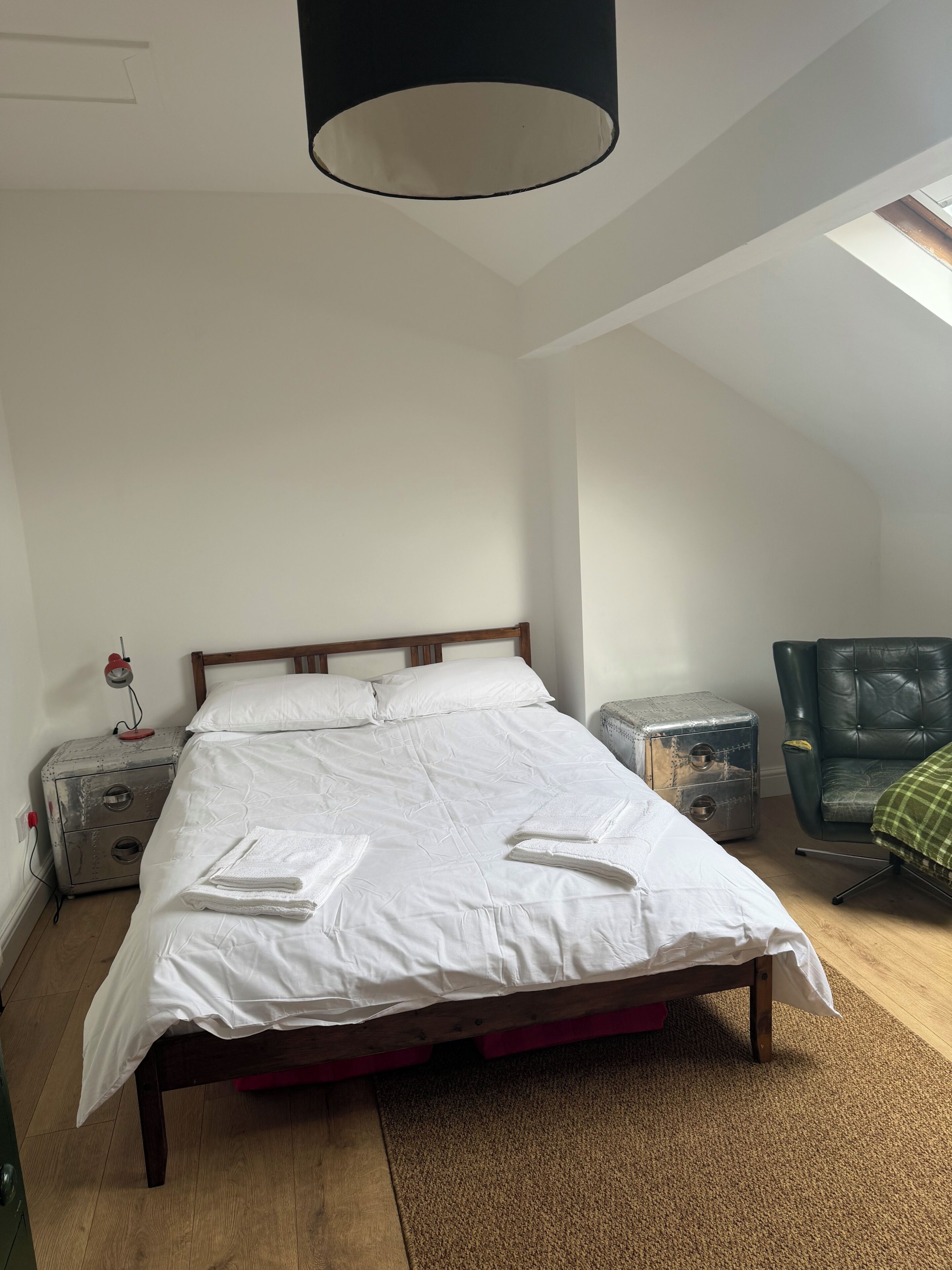 Un dormitorio con cama de madera, una mesita de noche pequeña, una silla verde y un tragaluz.