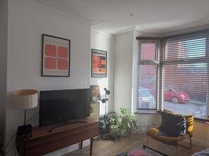 A living room with a flat-screen TV, a lamp, a chair, and two potted plants. - Boho Retro 4-bedroom house in charming Headingley for your Leeds stay (Leeds)