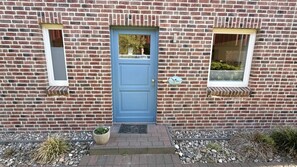 A brick building with a blue door, two windows, and a small garden area with rocks and plants. - Vacation apartment Möwe Bauernhof-Claussen (Fehmarn)
