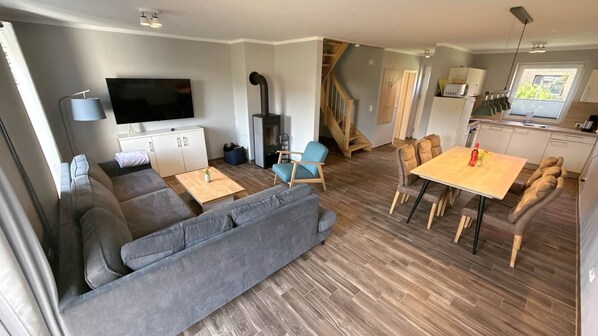 A modern living room with a sofa, TV, fireplace, dining area, and kitchen. - Vacation apartment Möwe Bauernhof-Claussen (Fehmarn)