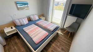 A bedroom with a bed, bedside tables, a TV, and a window with curtains. - Vacation apartment Muschel Bauernhof-Claussen (Fehmarn)