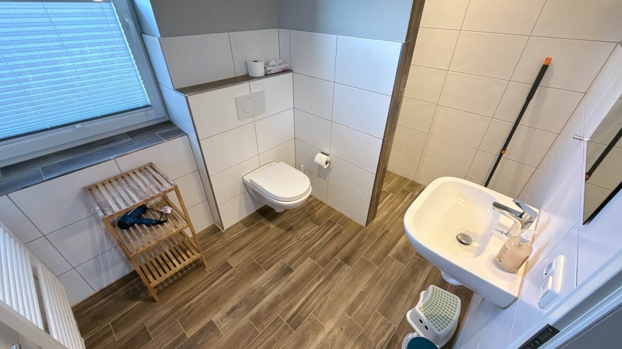 Une petite salle de bain avec des toilettes, un lavabo, un plancher en bois et une fenêtre avec des stores.