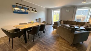 A modern living room with a dining table, sofas, and a large window. - Vacation apartment Welle Bauernhof-Claussen (Fehmarn)