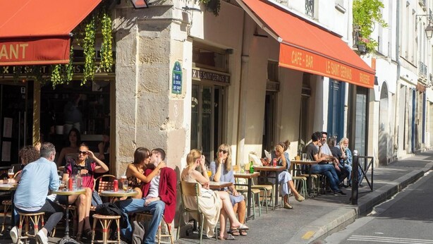 Terrasse d'un café-bar avec tables et chaises, donnant sur une rue et située devant un immeuble avec une enseigne.