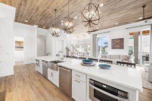 A modern kitchen with white cabinetry, stainless steel appliances, and a large island. There are pendant lights with a geometric design, and the ceiling has a wooden finish. - Huge Corner House 30A! Free Bikes/Walk to Beach! (Santa Rosa Beach)
