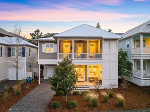 A two-story residential building with a front porch, a garage, and a small tree in the front yard.