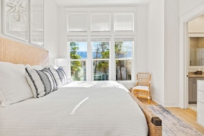 Ein geräumiges Schlafzimmer mit einem großen Bett, einem Korbstuhl und Blick auf Bäume durch das Fenster.