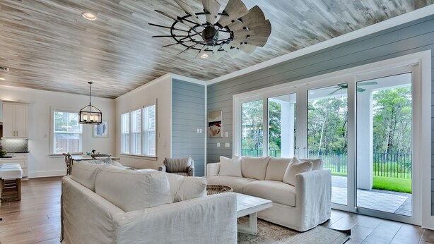 Un salón amplio con un sofá sectional blanco grande, una mesa de centro y un ventilador de techo. Amplias ventanas ofrecen vistas al exterior.