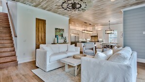 Un salón amplio con un sofá blanco, una mesa de centro y un comedor con una silla a rayas. Se ve una escalera de madera a la izquierda.