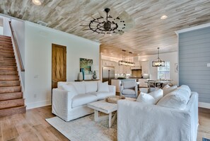 Ein geräumiges Wohnzimmer mit einem weißen Sofa, einem Couchtisch und einem Essbereich mit einem gestreiften Stuhl. Eine Holztreppe ist links im Bild zu sehen.