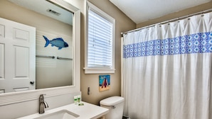 Een badkamer met een wit wastafel, toilet, een douchegordijn met blauw patroon, een sticker van een vis op de muur, en een raam met jaloezieën.