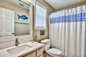 Ein Badezimmer mit einem weißen Waschbecken, einer Toilette, einem blauen Duschvorhang mit Muster, einem Fisch-Aufkleber an der Wand und einem Fenster mit Jalousien.