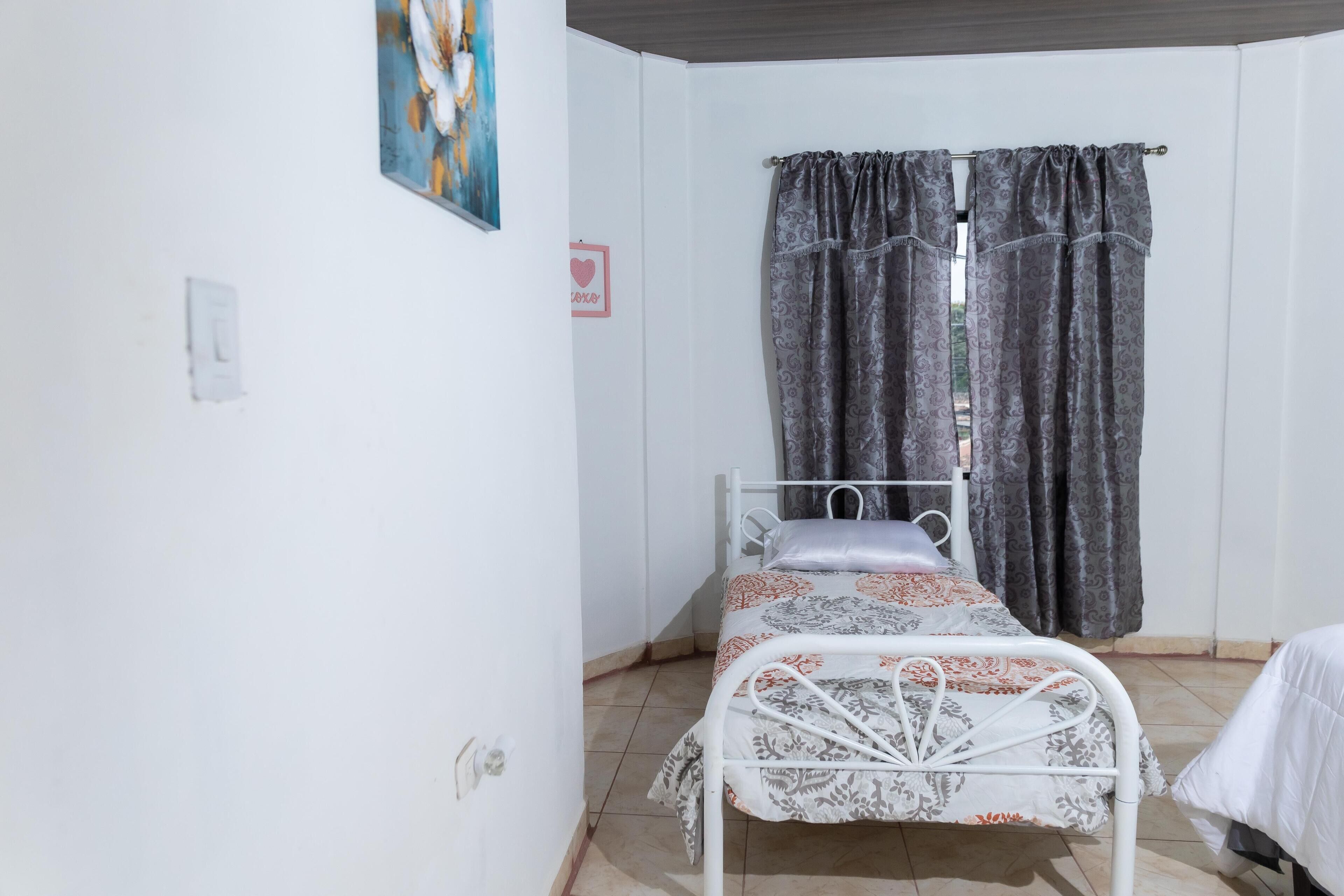 A single-bed room with a metal frame, a patterned bedspread, a window with patterned curtains, and a wall-mounted painting.