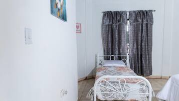 A single-bed room with a metal frame, a patterned bedspread, a window with patterned curtains, and a wall-mounted painting.