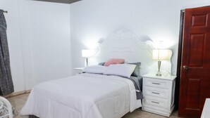A neatly made bed with white bedding, a nightstand with a lamp, a wooden door, and a white dresser with drawers. - La Casona Blanca (San Marcos)