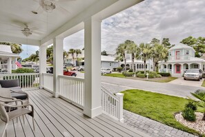 A covered porch with wicker furniture, a view of a residential street with palm trees, and houses with a red door. - 30A Family House! Free Bikes. Walk to Coffee/Dinner! (Santa Rosa Beach)