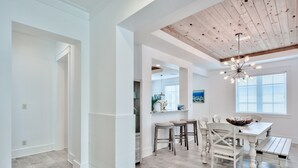 Une cuisine spacieuse avec un plafond en bois, un coin repas avec une table et des chaises blanches, et une grande fenêtre équipée de stores.