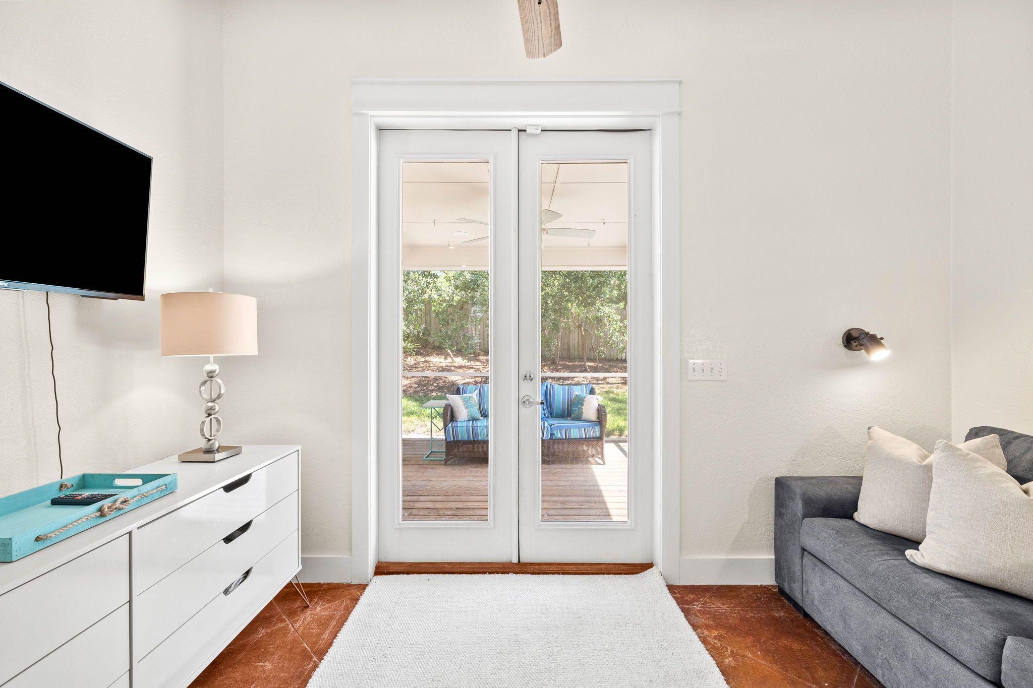 A room with a white wall, a flat-screen TV mounted above a white dresser, a grey sofa with cushions, and a sliding glass door leading to an outdoor deck with a blue chair and a table.