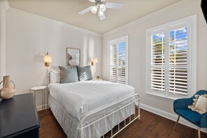 Une chambre à coucher comprenant un lit, des tables de chevet, une chaise, un ventilateur de plafond et des fenêtres équipées de stores.