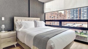 Een moderne slaapkamer met een groot raam, een bed met witte lakens, een nachtkastje en een lamp.
