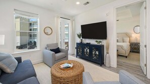 A spacious living room with a large window, a ceiling fan, a flat-screen TV, a sofa, a chair, and a small round table. - Gulf Place 30A! Walk to Beach. (SANTA ROSA BEACH)