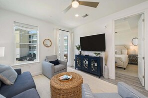 A spacious living room with a large window, a ceiling fan, a flat-screen TV, a sofa, a chair, and a small round table. - Gulf Place 30A! Walk to Beach. (SANTA ROSA BEACH)