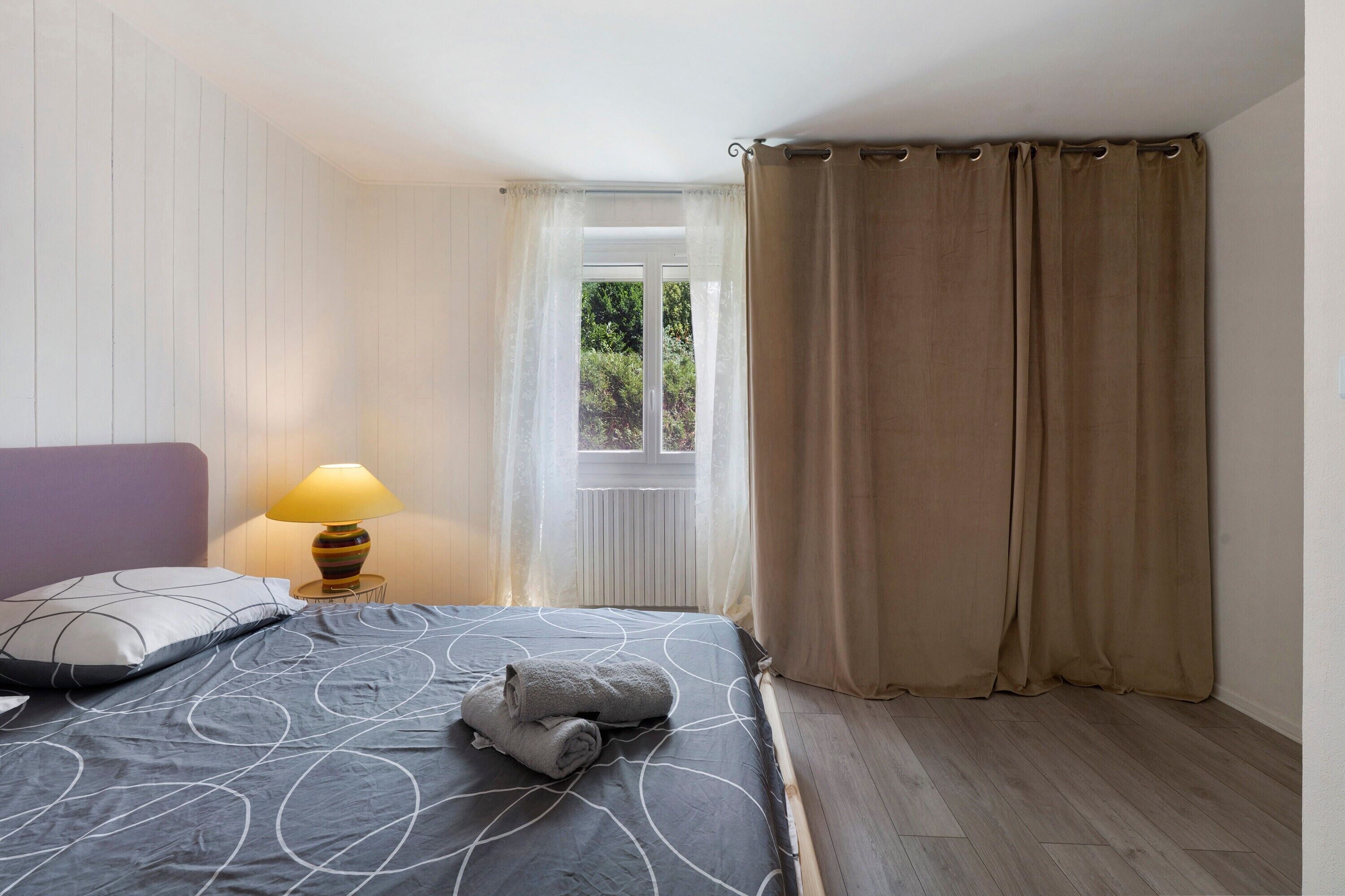 A bedroom with a bed, a nightstand with a lamp, a window with curtains, and a view of greenery outside.