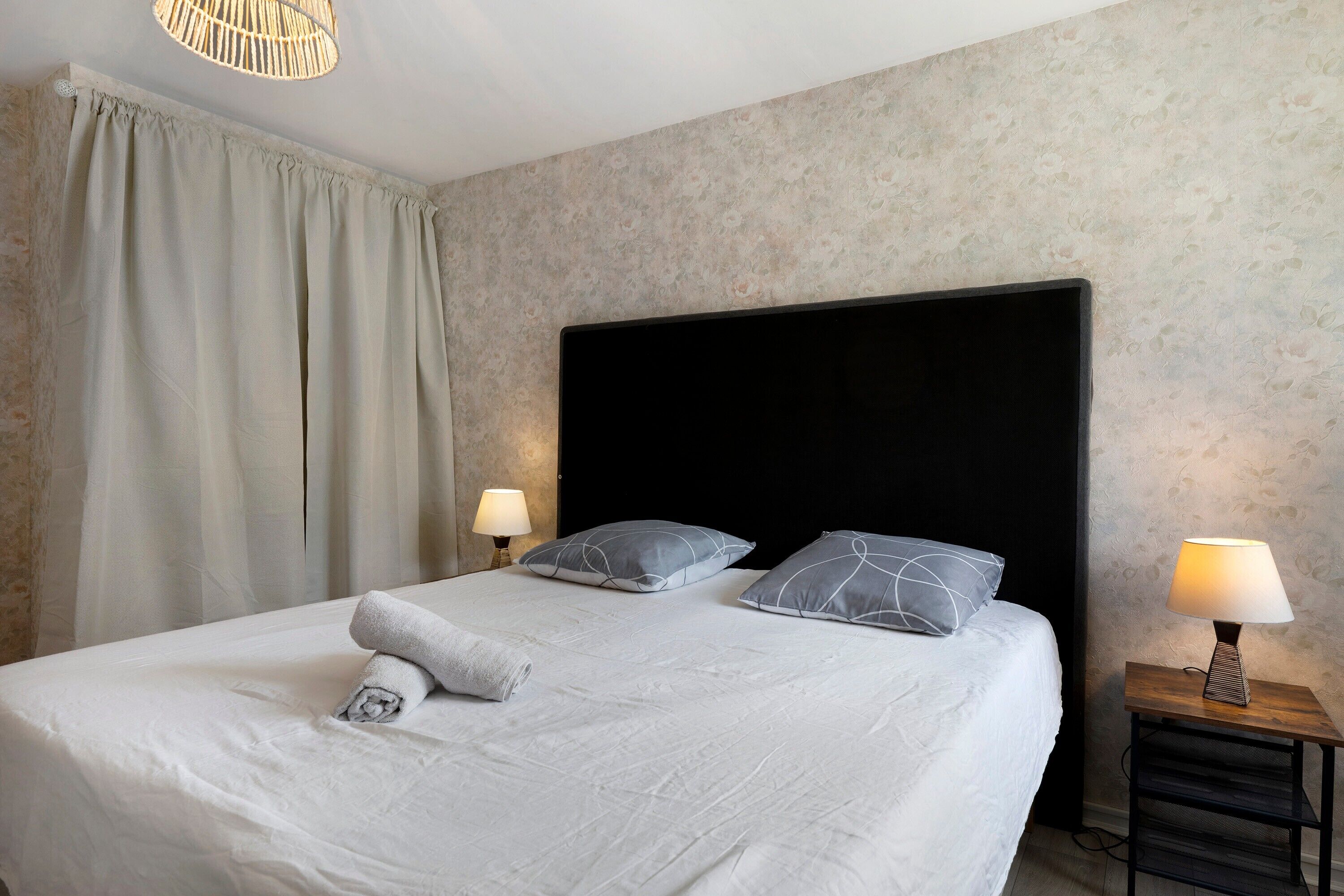 Ein ordentlich bezogenes Bett mit Kissen und einem zusammengerollten Handtuch, eine Nachttischlampe und eine Wand mit einem Blumenmuster.