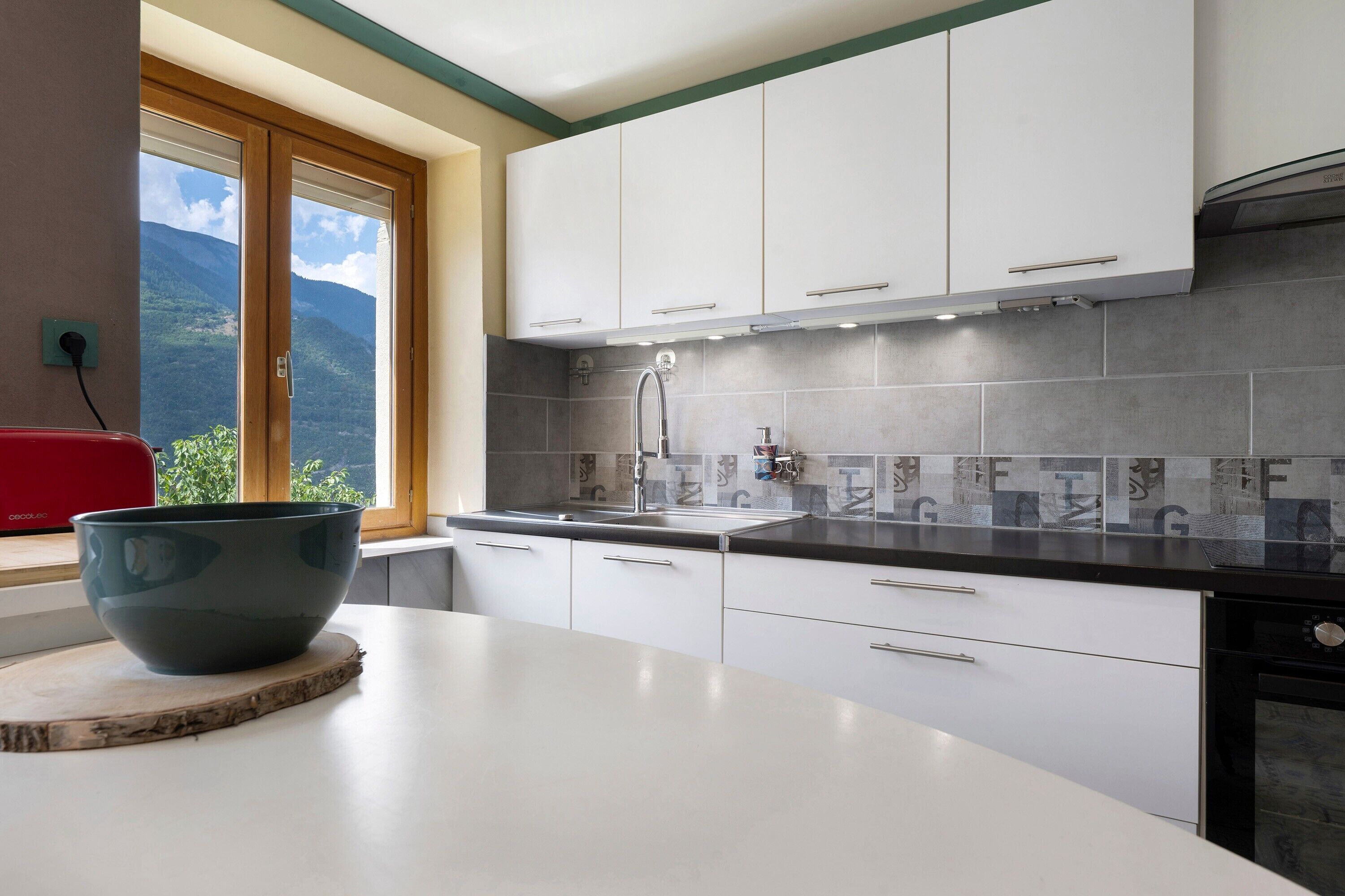 Une cuisine moderne avec des meubles blancs, un pare-feu en carreaux gris et une grande fenêtre donnant sur les montagnes.