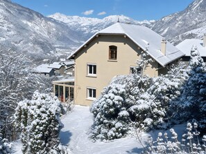 A house with a sloped roof, surrounded by snow-covered trees and mountains in the background.