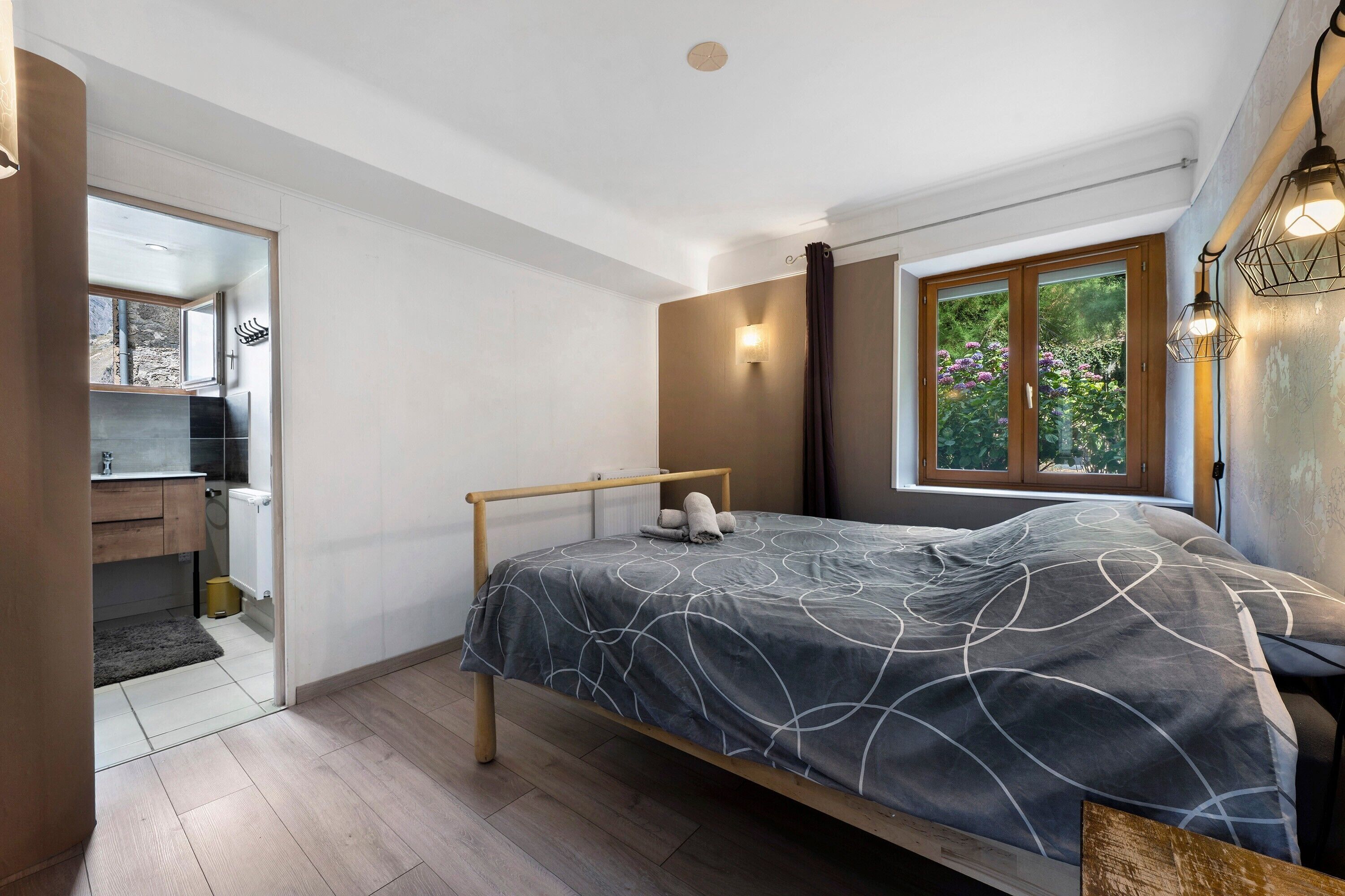 Une chambre à coucher avec un lit, une fenêtre avec des rideaux, un radiateur et une salle de bain visible à travers une porte ouverte.