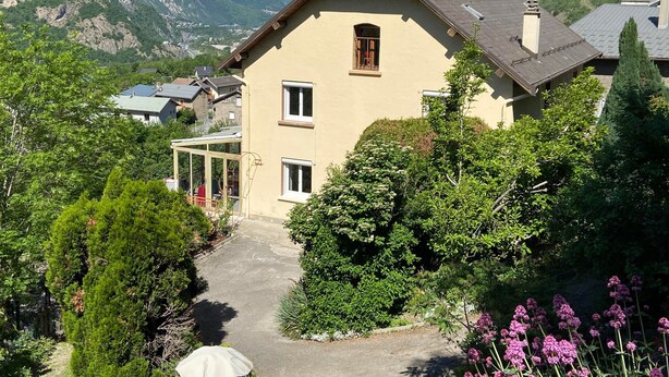 A house with a sloped roof, a visible window, and a garden with various plants and flowers.